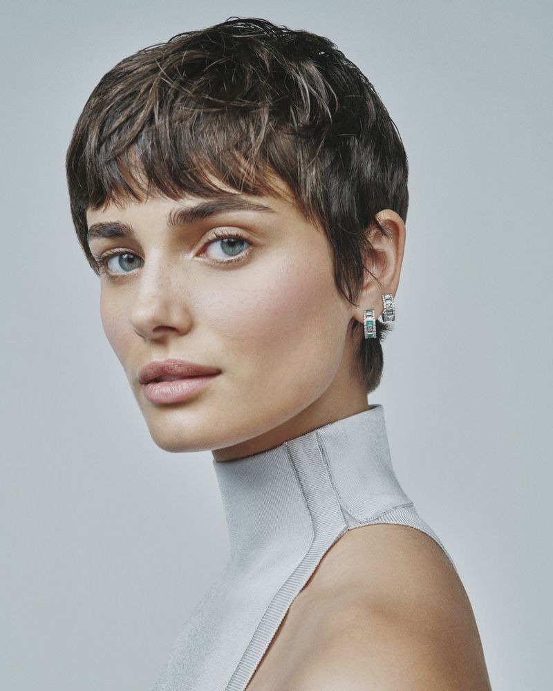 Close-up portrait of a woman with short brown hair wearing silver hoop earrings against a neutral grey background in a beauty shot.