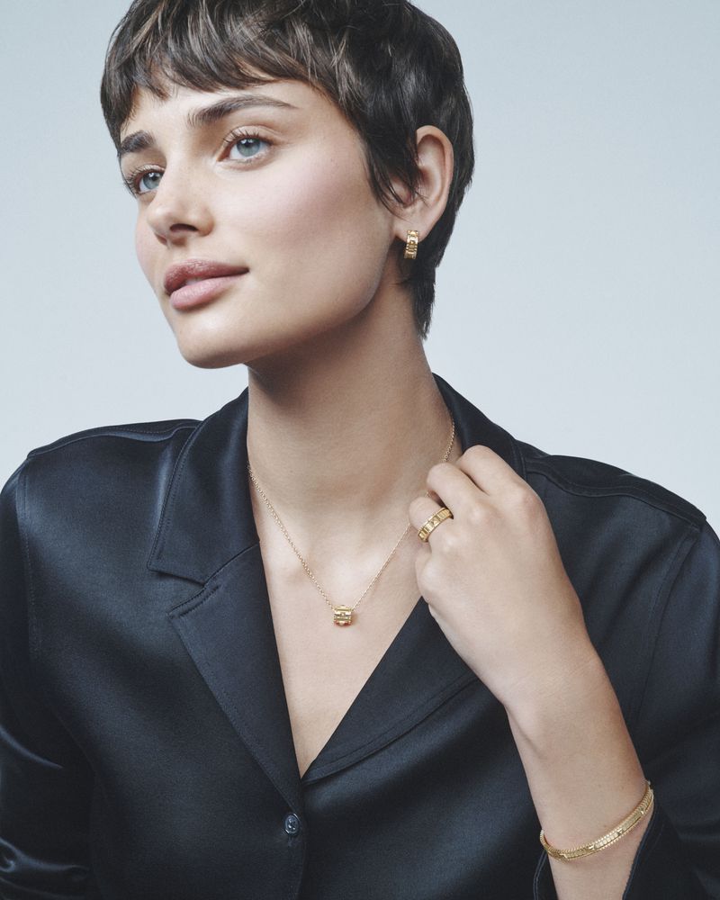 Portrait of a woman wearing gold jewelry including a necklace, ring, bracelet and earrings, with a soft light blue background.