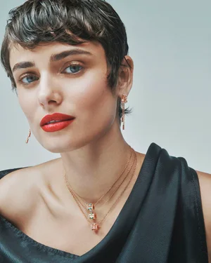 Close-up portrait of model Taylor Hill wearing layered rose gold necklaces, drop earrings, and red lipstick against a grey background.
