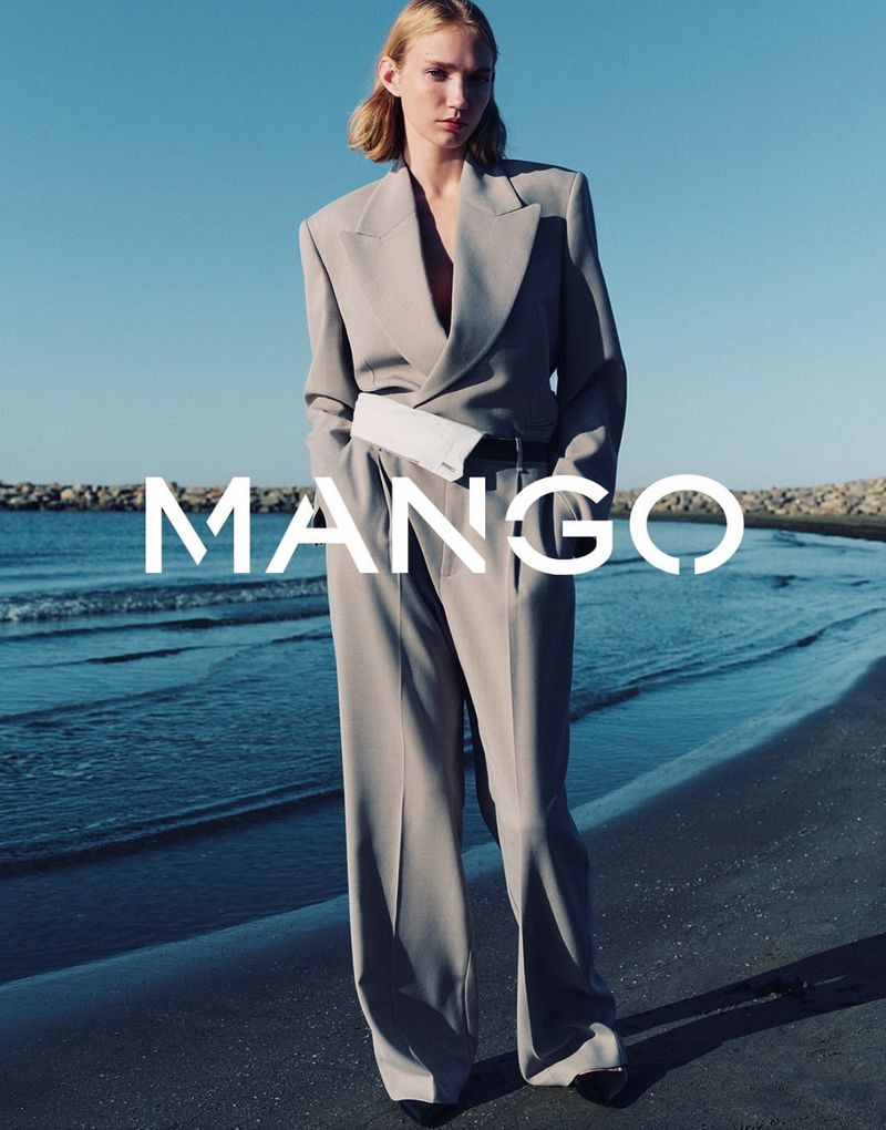Blonde woman in grey business suit and white belt bag poses on beach with ocean and rocks; fashion editorial style, Mango brand signage.