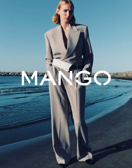 Blonde woman in grey business suit and white belt bag poses on beach with ocean and rocks; fashion editorial style, Mango brand signage.