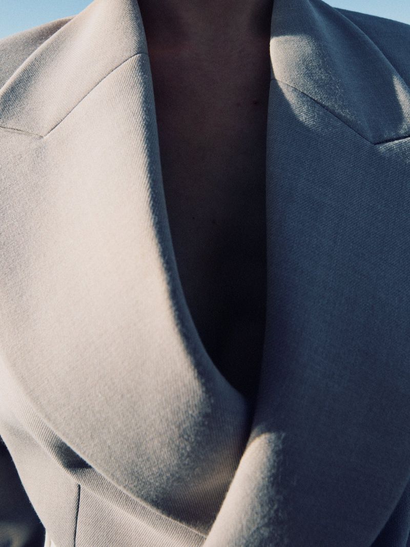 A close-up shot of a person wearing a light-colored coat, emphasizing the textures and style of the garment against a bright sky.