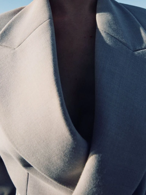 A close-up shot of a person wearing a light-colored coat, emphasizing the textures and style of the garment against a bright sky.