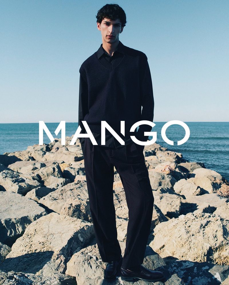 Dark-haired male model in a long-sleeve shirt and slacks stands on rocks by the ocean in a fashion shot by Mango. 