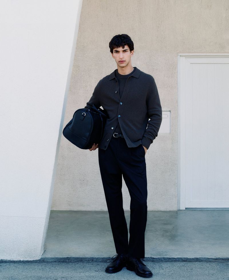 Man wearing a dark cardigan and trousers holding a black duffel bag standing outside a building for Mango Fall Winter 24 campaign