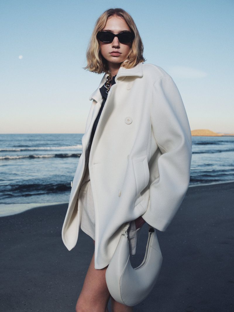 Fashion photography featuring a fair-skinned young adult woman in a white coat on a sandy beach with ocean and clear sky backdrop.