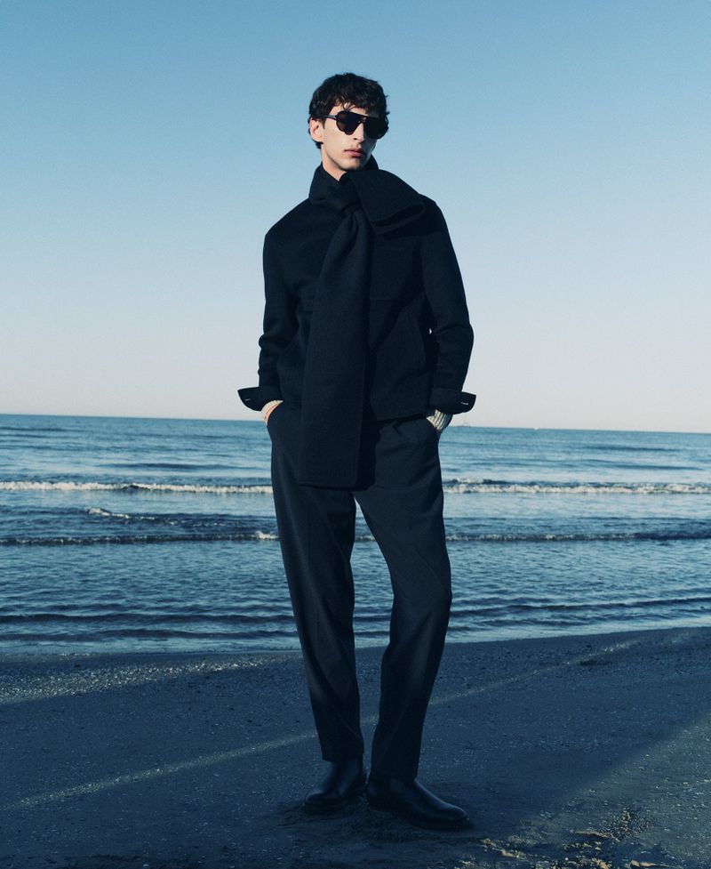 Model wearing dark sunglasses and a black sweater standing on a sandy beach by the ocean under a clear sky.