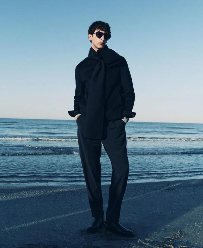 Model wearing dark sunglasses and a black sweater standing on a sandy beach by the ocean under a clear sky.