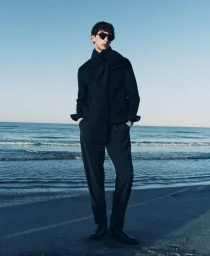Model wearing dark sunglasses and a black sweater standing on a sandy beach by the ocean under a clear sky.