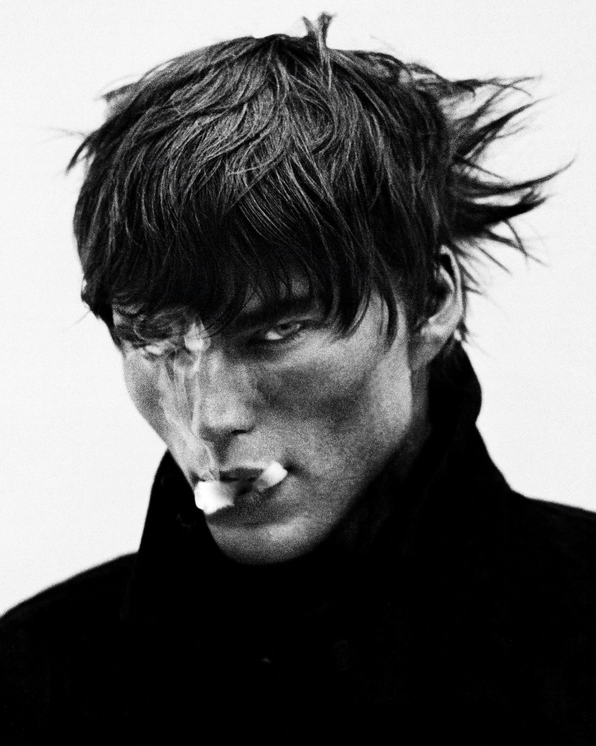 Monochrome portrait of a young adult man with dark messy hair, smoking and exhaling smoke. He is wearing a black coat.