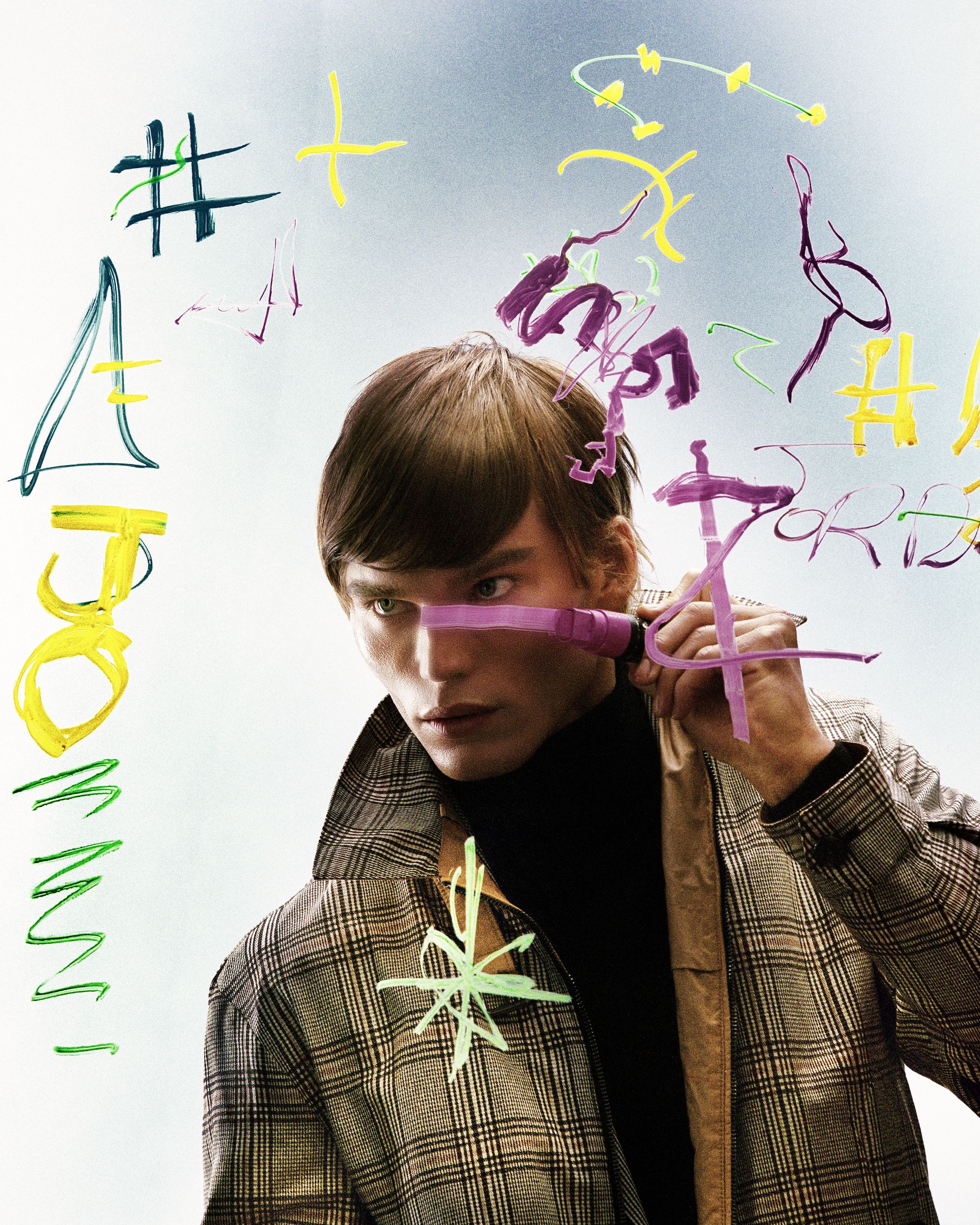 Editorial fashion shot of a fair-skinned young man with brown hair wearing a plaid coat and holding a pink paintbrush in an indoor environment.