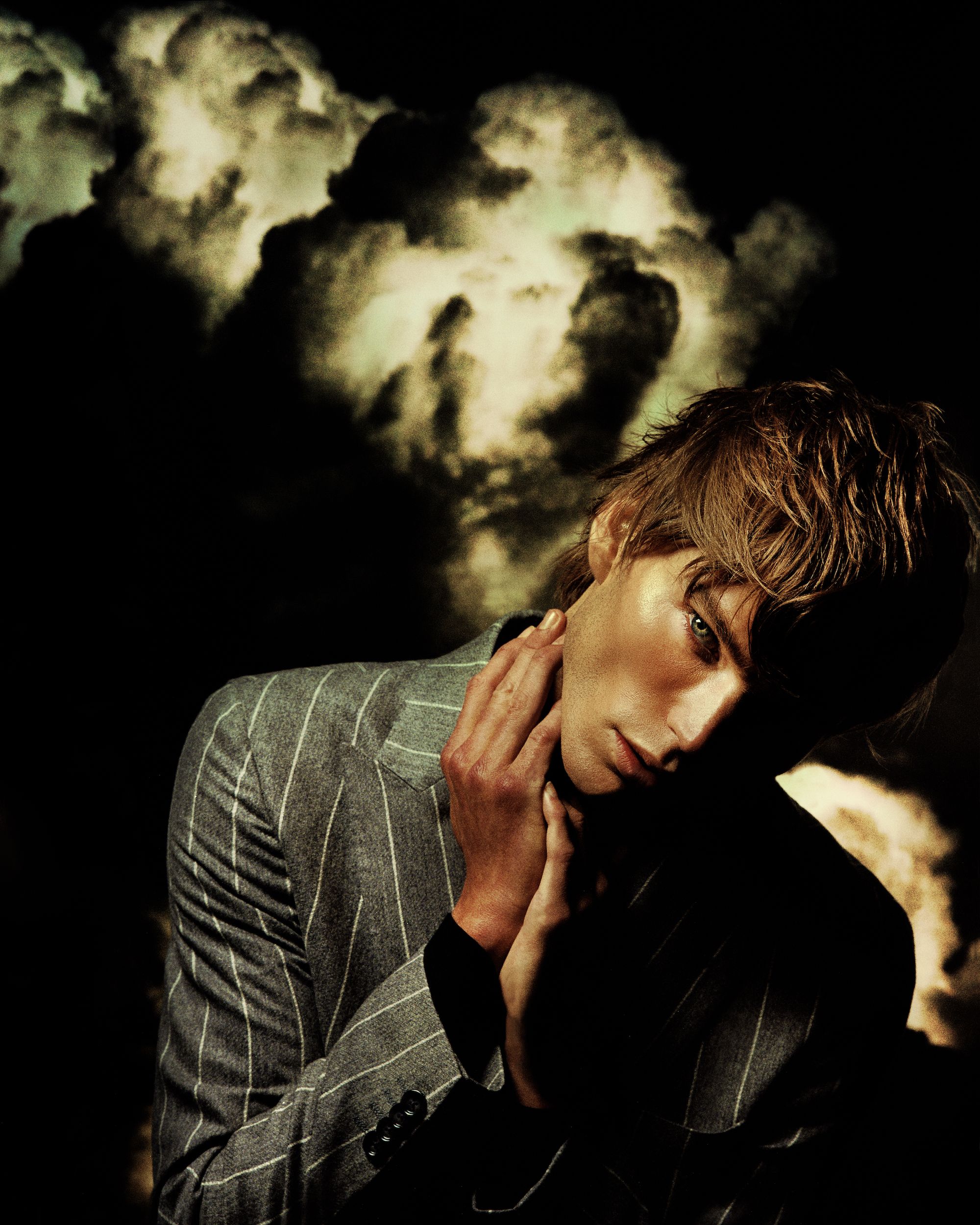 Portrait of a young adult male in a pinstripe blazer, hands near his neck, posing against a dark background resembling stormy clouds.