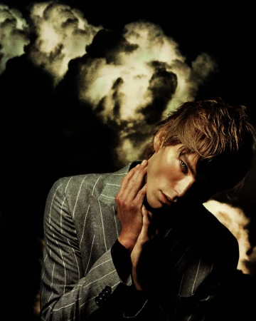 Portrait of a young adult male in a pinstripe blazer, hands near his neck, posing against a dark background resembling stormy clouds.