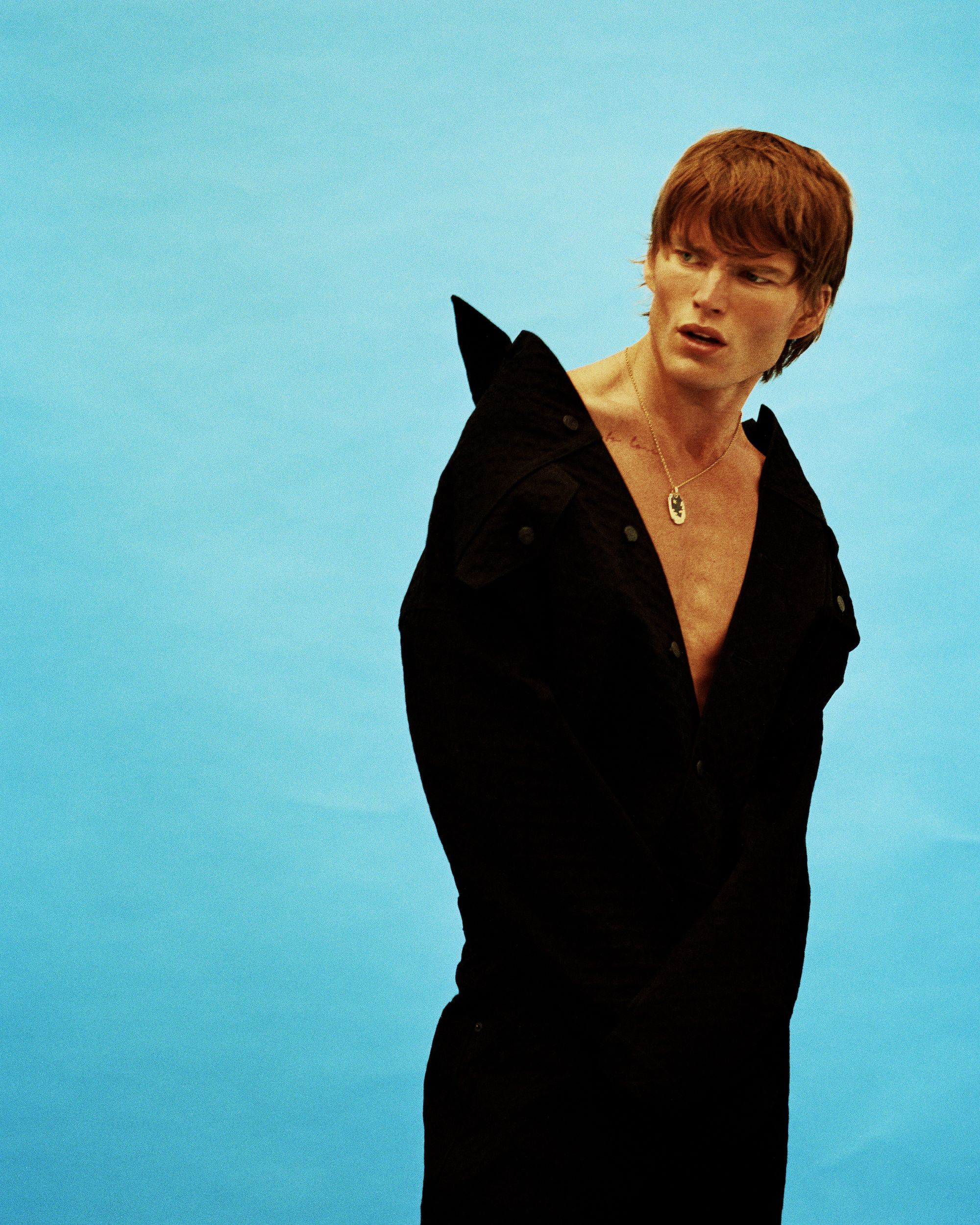 Attractive ginger male model in a partially unbuttoned black shirt, looking sideways against a bright blue backdrop in a studio setting.