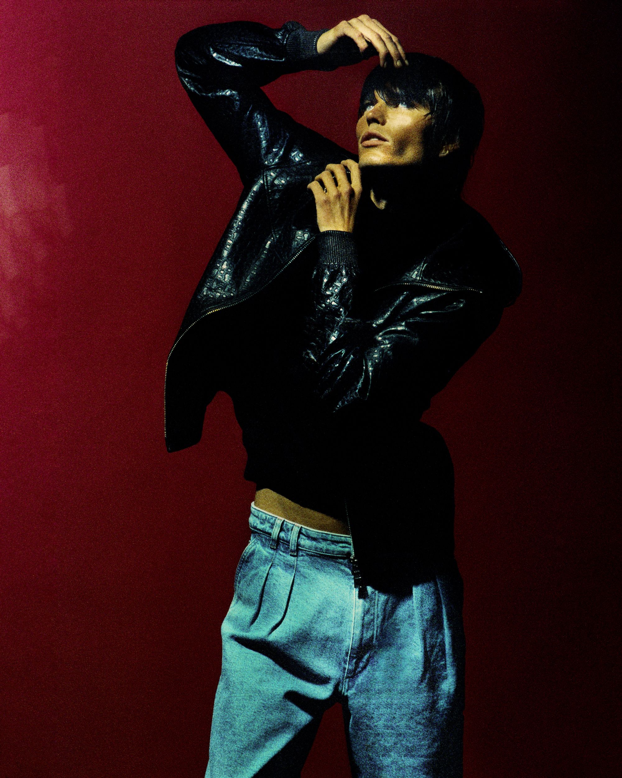 Model in a black jacket and blue denim trousers posing against a dark red background with dramatic lighting.