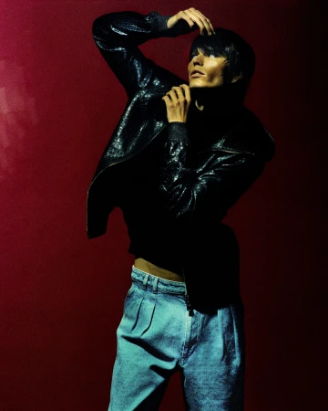 Model in a black jacket and blue denim trousers posing against a dark red background with dramatic lighting.