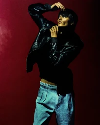 Model in a black jacket and blue denim trousers posing against a dark red background with dramatic lighting.