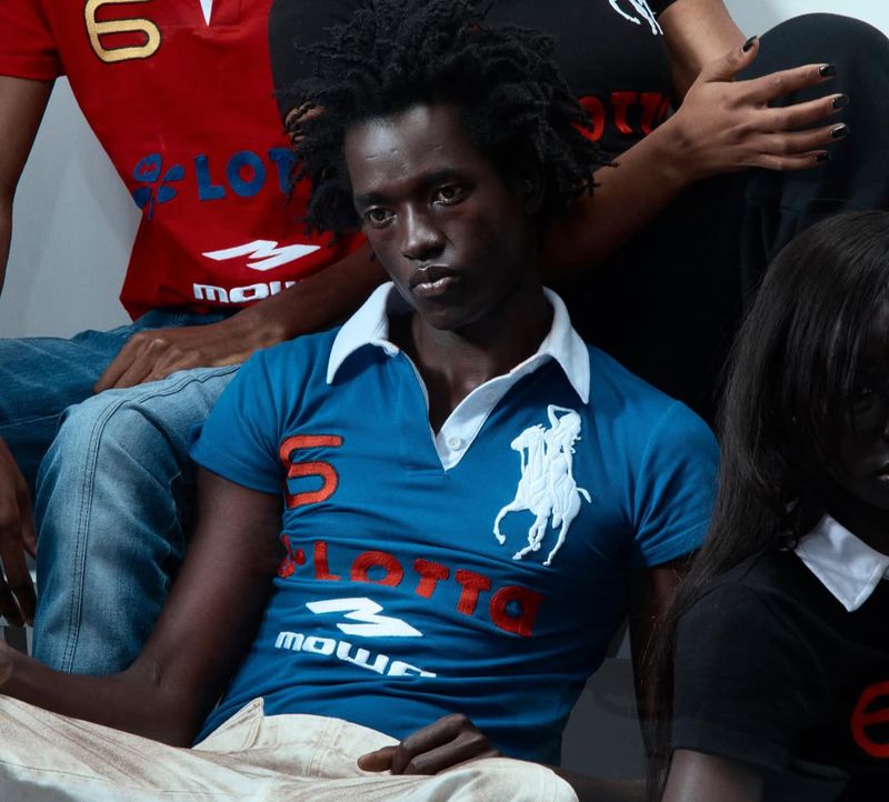 Group shot of young adults wearing polo shirts with logos, posing indoors. Blue, red, and black shirts are visible with a white wall backdrop.