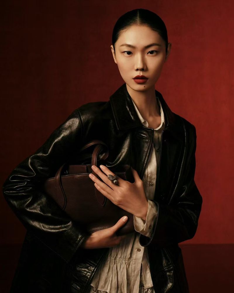 A fashionable Asian woman in a black leather jacket poses indoors with a brown handbag against a solid red backdrop.