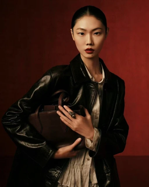 A fashionable Asian woman in a black leather jacket poses indoors with a brown handbag against a solid red backdrop.