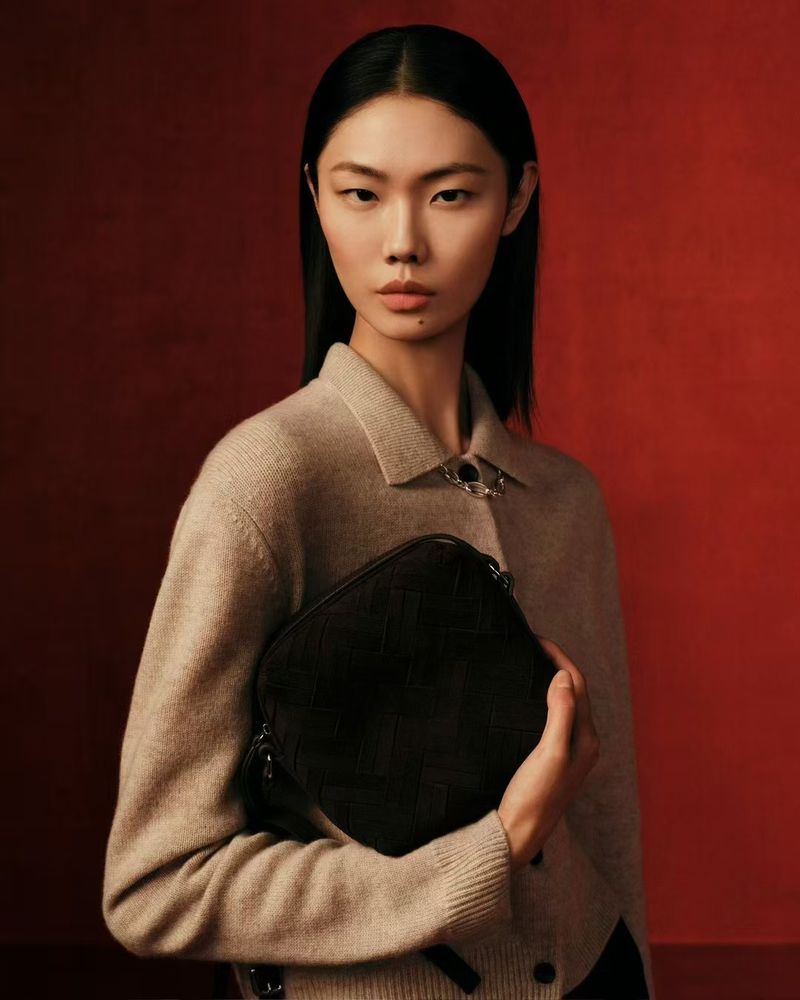 Asian woman with black hair posing indoors against a red background, wearing a beige cardigan and holding a black leather bag.