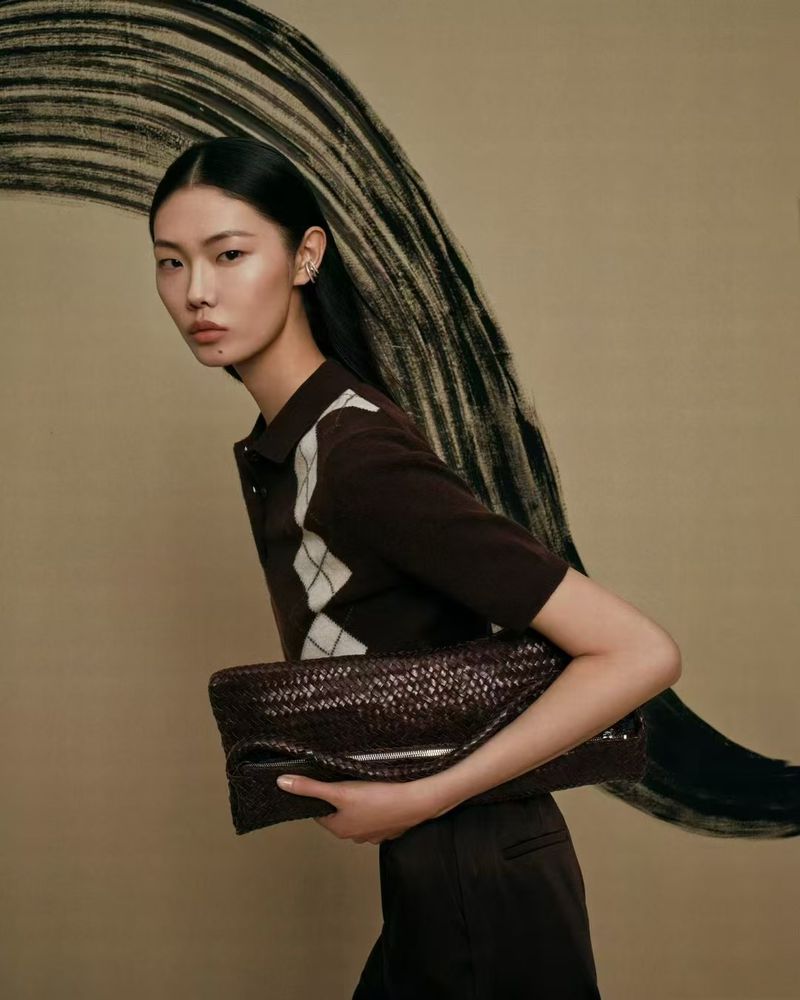 Woman in brown argyle shirt holding woven leather clutch bag against neutral backdrop with black brushstroke design in a studio setting.