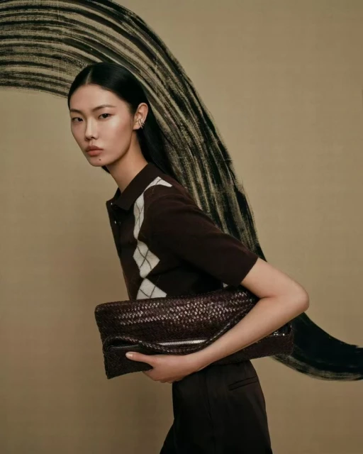 Woman in brown argyle shirt holding woven leather clutch bag against neutral backdrop with black brushstroke design in a studio setting.