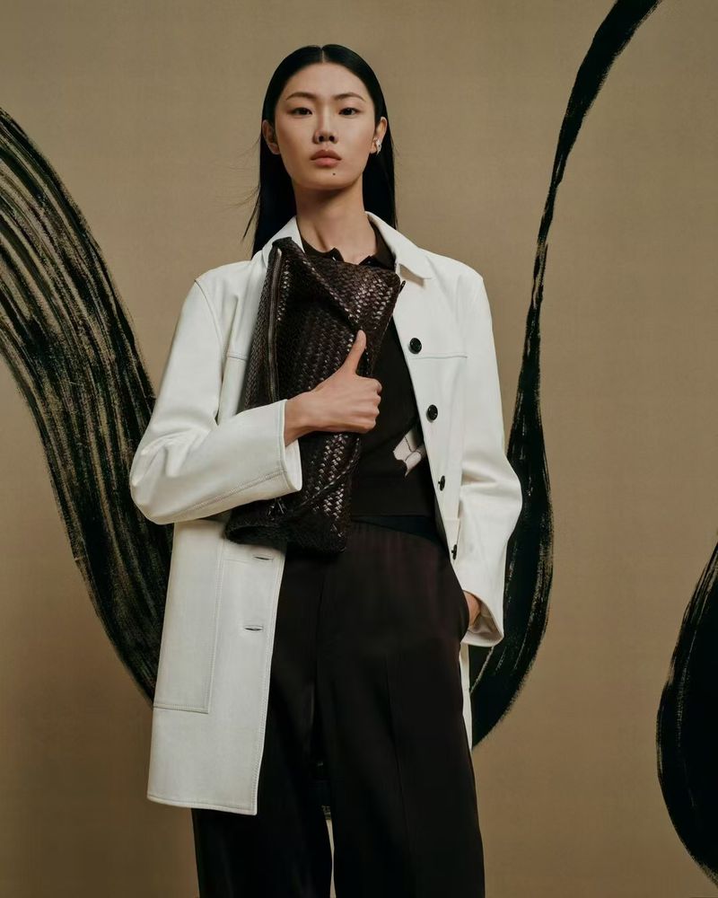 Asian woman in a white jacket and dark brown pants holds a woven handbag, posing in a studio with abstract black brushstrokes.