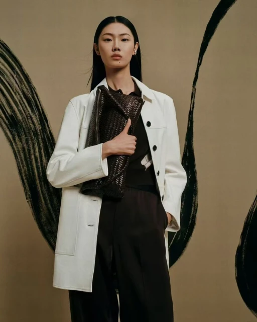 Asian woman in a white jacket and dark brown pants holds a woven handbag, posing in a studio with abstract black brushstrokes.