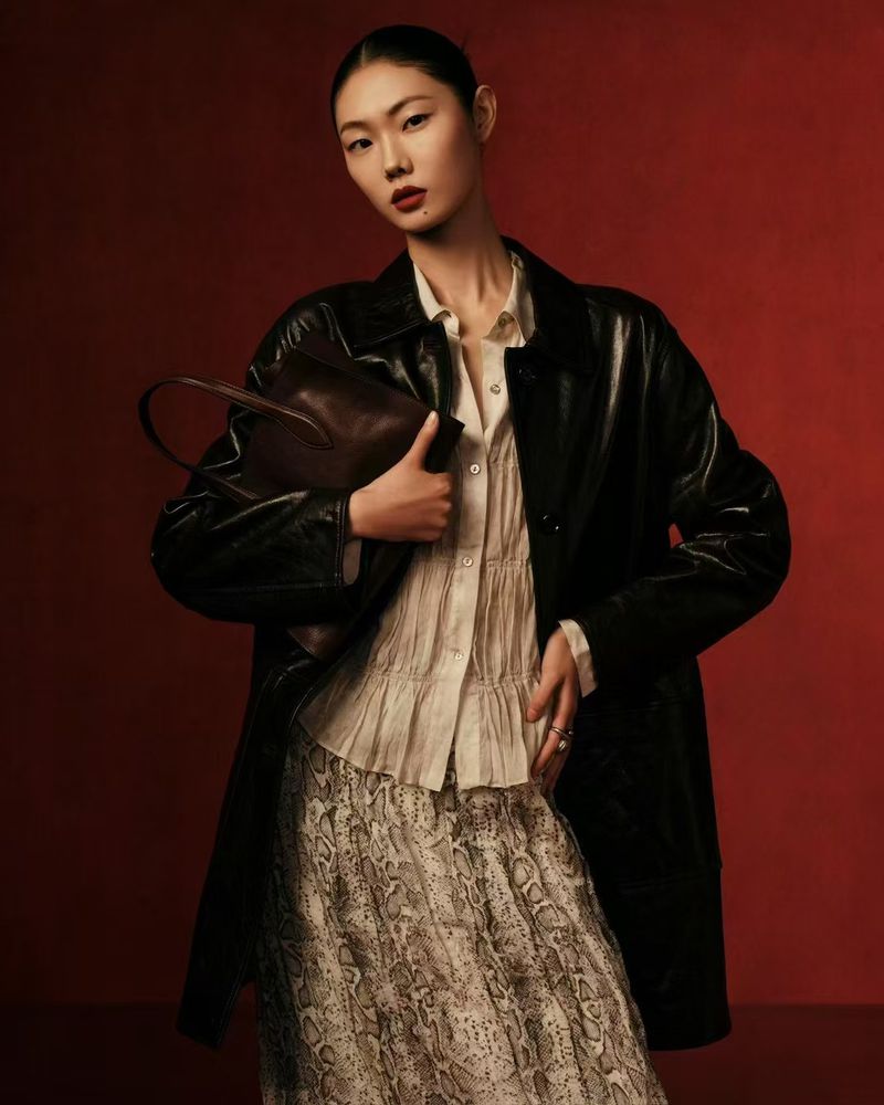A stylish Asian woman poses in a studio, wearing a black jacket, printed skirt, and carrying a leather handbag against a red backdrop.