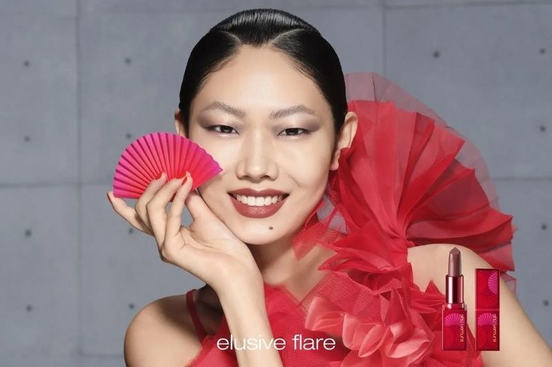 Asian female model in a red dress smiles holding a red fan, next to a red lipstick tube in a beauty advertisement against a gray background.