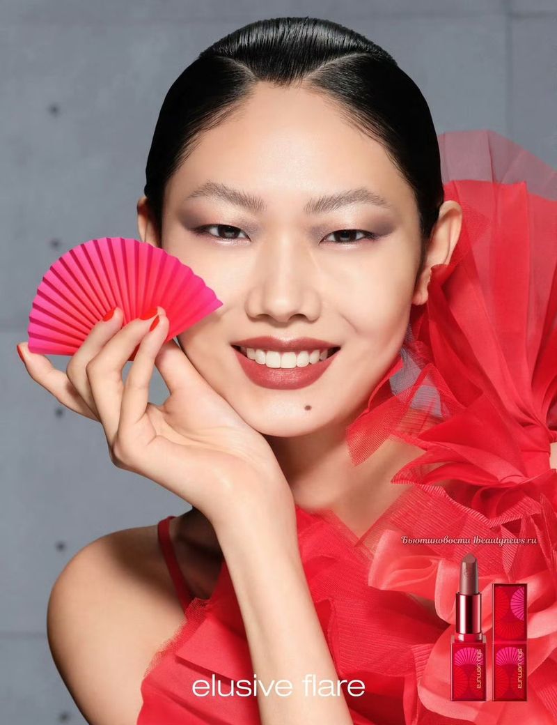 Asian woman poses with a pink fan and red dress in a studio setting, showcasing elegant makeup and a lipstick product for beauty advertising.