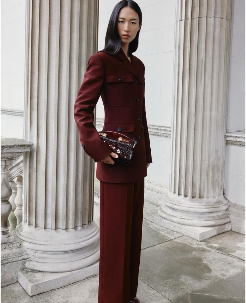 Asian woman in a burgundy suit with wide-leg pants, holding a dark, ornate handbag, posing against a backdrop of stone columns.