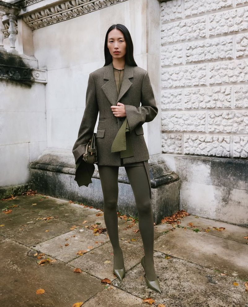 An Asian woman poses on a city sidewalk in a stylish olive green jacket, matching tights and heels, and carrying a purse.