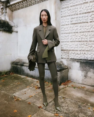 An Asian woman poses on a city sidewalk in a stylish olive green jacket, matching tights and heels, and carrying a purse.