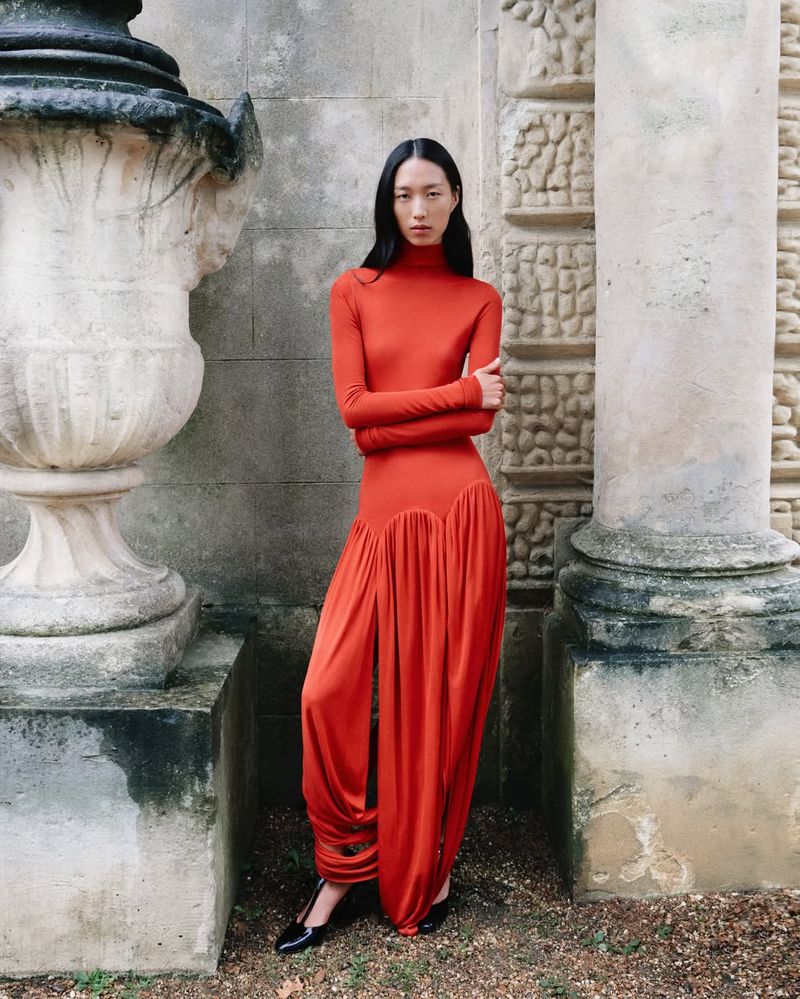 Asian woman in a long-sleeved orange gown poses by architectural stone wall with classic urn in daylight for a fashion editorial.
