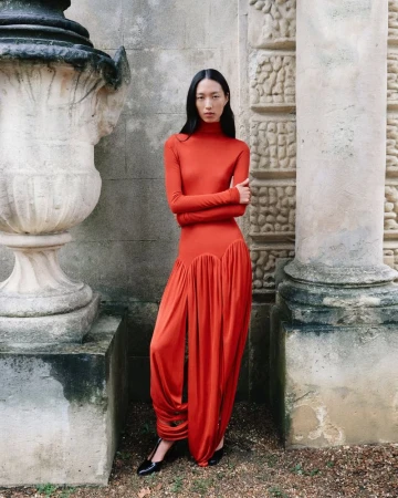 Asian woman in a long-sleeved orange gown poses by architectural stone wall with classic urn in daylight for a fashion editorial.