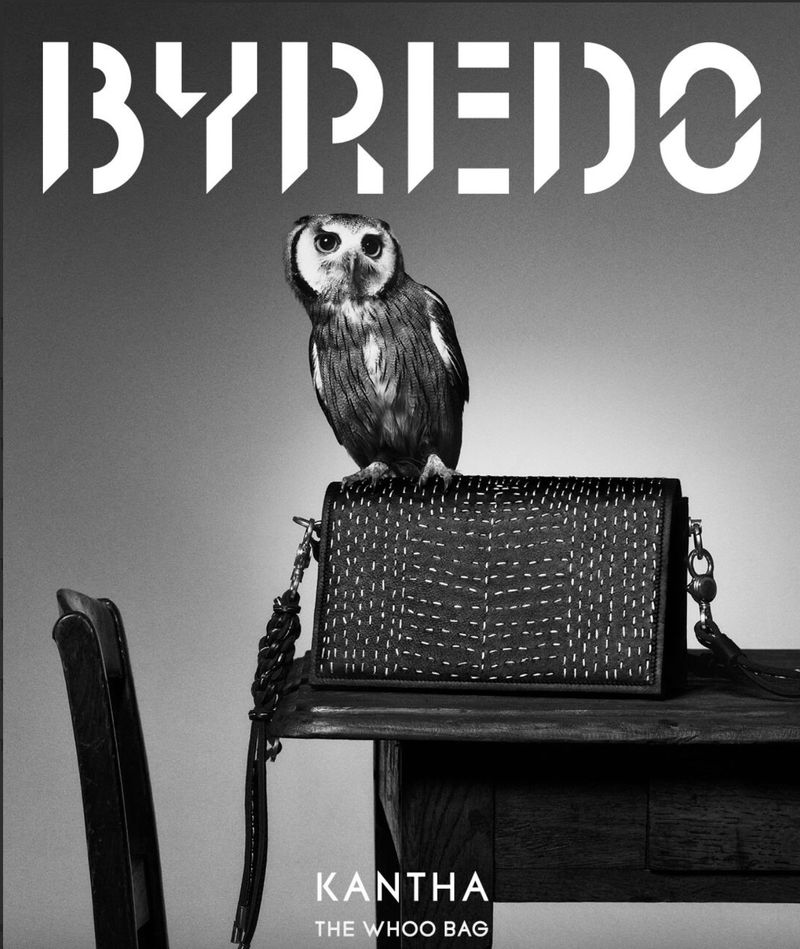 Black and white shot of a Byredo purse with an owl perched on top, sitting on a wooden table next to a black chair.
