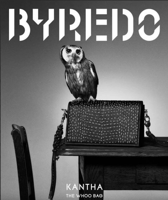 Black and white shot of a Byredo purse with an owl perched on top, sitting on a wooden table next to a black chair.