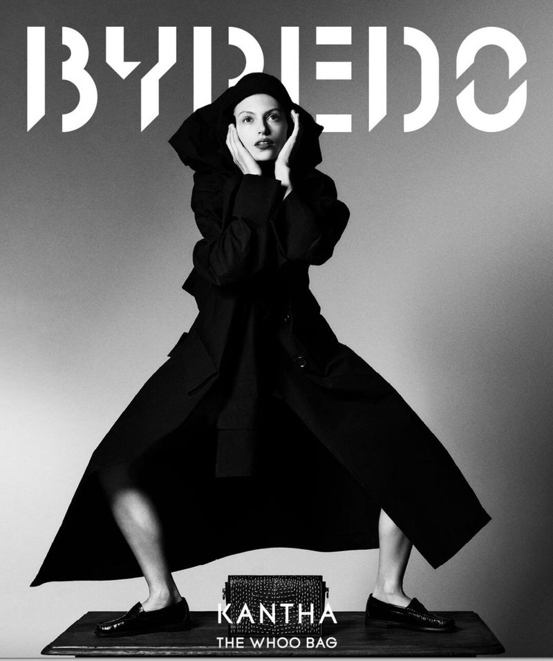 Model wearing a black hooded coat poses with hands framing her face over a textured black bag on a table against a grey gradient background
