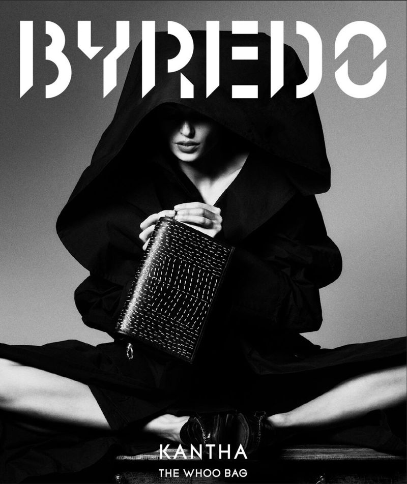 Fashion shot in black and white features a woman in dark hooded clothing holding a textured black handbag, seated in a dark photo studio.