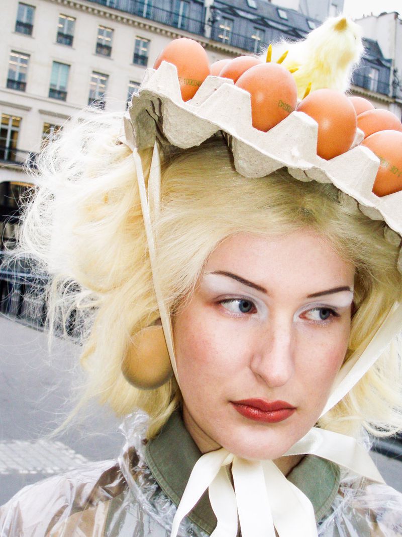 Woman with blonde wig wears egg carton hat and translucent coat in urban setting. Eggs are visible on the hat and as earrings.