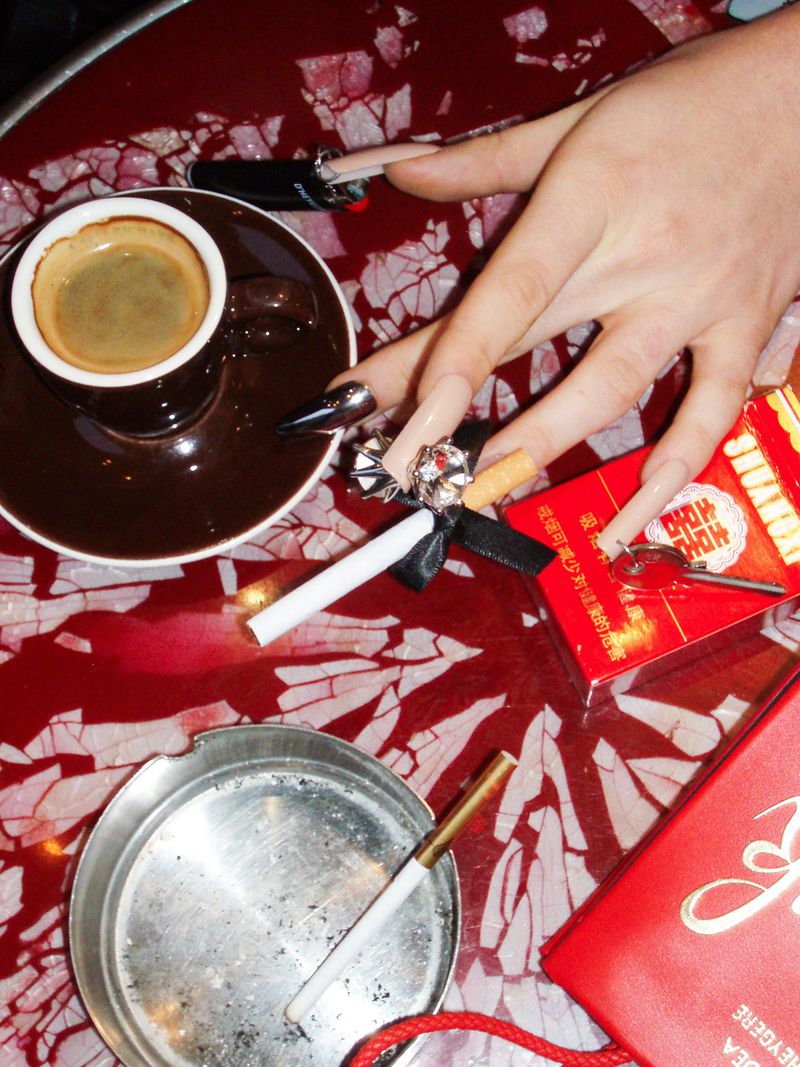 Overhead shot of a hand with long nails holding a cigarette, with an espresso, cigarettes, and ashtray on a red patterned table.