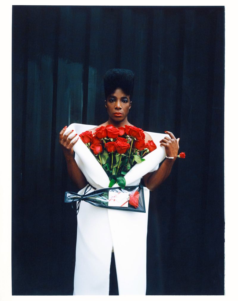 Woman in white dress holding a bouquet of red roses against a black backdrop, jewelry is visible on her wrist.