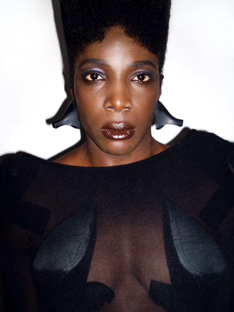 Close-up portrait of a dark-skinned woman with a high-top fade, wearing large geometric earrings and a black semi-sheer top against a white backdrop.