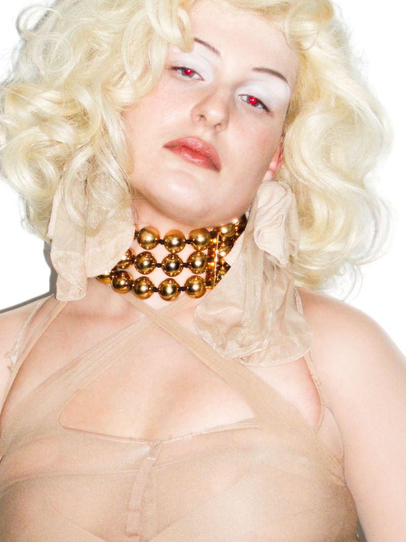 Portrait of a woman with blonde curly hair, dramatic white makeup, a gold necklace, and a sheer, nude-colored garment against a white background.