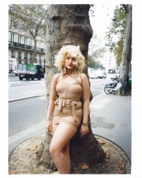 Model with blonde hair wearing a beige dress leaning against a tree on a city street
