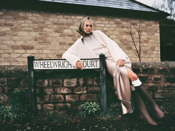 Fashionable woman in headscarf leans on Wheelwright Court sign. She wears cream sweater, skirt, boots in outdoor setting with brick building.