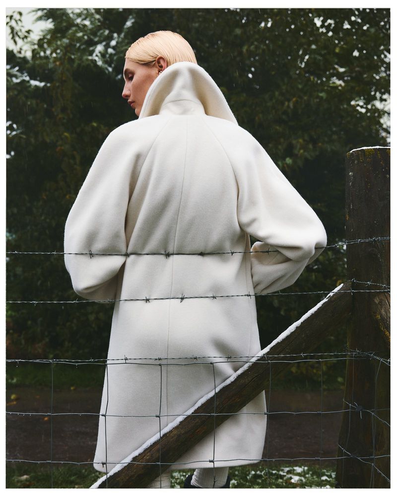 A blonde-haired model in a cream overcoat poses by a snow-covered wire fence with an out-of-focus green forest in the background.