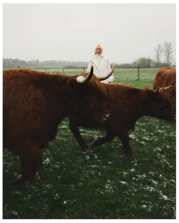 A woman in a white coat stands behind a herd of brown Highland cattle in a field on a cloudy day, blending fashion and nature.